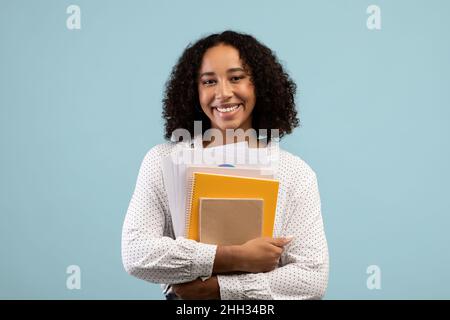Porträt einer jungen schwarzen Studentin mit Notizbüchern, die vor der Kamera posieren und auf blauem Studiohintergrund lächeln Stockfoto