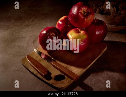 Apfelbrett. Äpfel und ein Messer stehen auf einem Schneidebrett und die ganze Komposition hat einen dunklen Hintergrund. Stockfoto