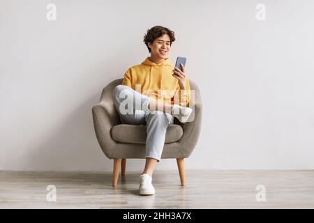 Hübscher junger Asiatischer, der Handy benutzt, im Internet oder in sozialen Medien surft, im Sessel sitzt und die neue mobile App überprüft Stockfoto