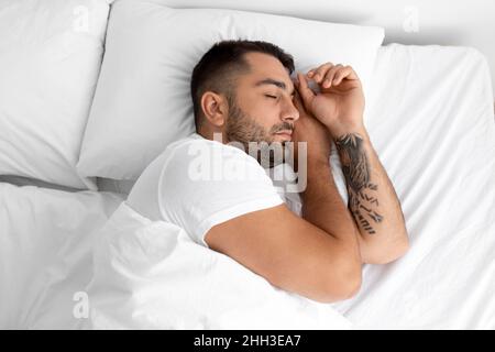 Ruhiger, tausendjähriger, hübscher Typ mit Bart schläft auf weißem, bequemen Bett auf weichem Kissen im Schlafzimmer, Blick von oben Stockfoto