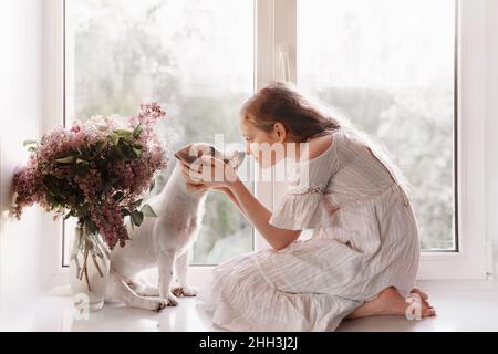 Nettes kleines Mädchen schaut in die Augen eines Hundes. Zwei Freunde sitzen am Fenster. Freundschaft, Fürsorge, glückliches Kindheitskonzept. Stockfoto