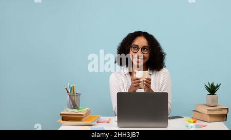 Glückliche schwarze Frau, die mit einem Laptop und Büchern am Schreibtisch sitzt, Kaffee trinkt, während des Online-Studiums oder der Arbeit Pause macht Stockfoto