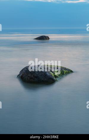 Blaue Stunde nach dem Sonnenuntergang über der felsigen Ostsee kosten. Kleine Steine und große Felsbrocken im Meer. Langzeitbelichtung Stockfoto