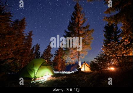 Nachtcamping in den Bergen. Drei beleuchtete Zelte, die auf Rasen unter Bäumen aufgestellt sind, leuchten rechts von den Zelten aus Flammen des Lagerfeuers. Campingplatz unter wunderschönem Sternenhimmel. Stockfoto