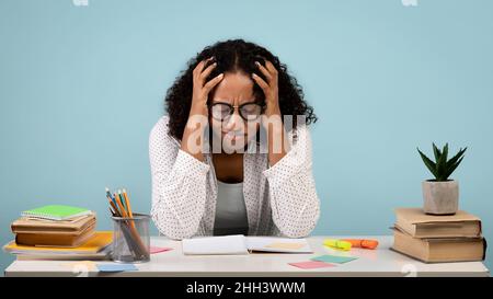 Müde afroamerikanische Studentin weint über Bücher und Notizbücher und studiert für schwierige Prüfung auf blauem Hintergrund Stockfoto