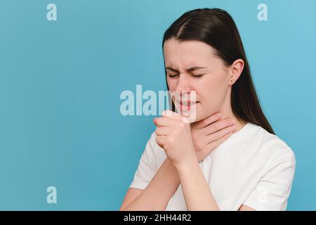 Ungesunde weibliche stehend isoliert auf blauem Hintergrund bedeckt ihren Mund mit der Hand, während sie hustet, leidet an saisonaler Grippe oder chronischer Bronchitis Stockfoto