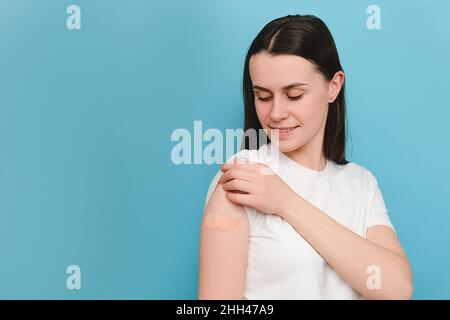 Porträt einer jungen kaukasischen Frau, die nach der Verabreichung einer Impfdosis im Studio mit CO auf blauem Hintergrund isoliert auf ihren Arm mit Pfandband schaut Stockfoto