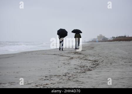 Skurrile, urige Paare, die an einem regnerischen Nachmittag an einem einsamen Strand mit Sonnenschirmen spazieren gehen Stockfoto