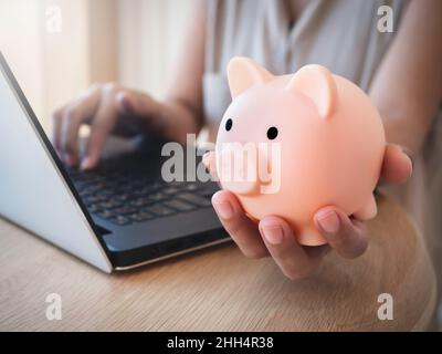 Geldbox zur Hand. Die Hand hält das pinke Sparschwein, während man den Laptop auf dem Schreibtisch benutzt. Geld sparen Reichtum und Finanzkonzept, Geschäft, online b Stockfoto