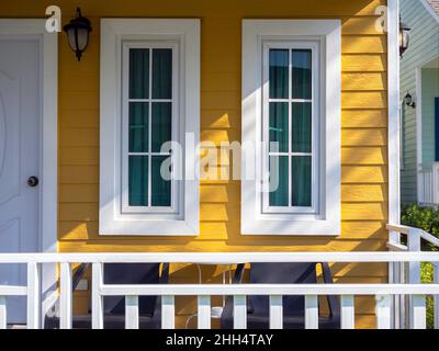 Zwei weiße Hausfenster mit Glas, grüner Vorhang Dekoration auf dem gelben Holz Bewohner in der Nähe der Haustür und Terrasse, Blick von draußen. Stockfoto