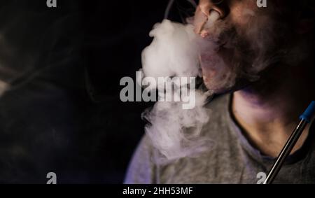 Mann Raucher, atmet Rauch aus. Dampf aus einer elektronischen Zigarette. Porträt eines jungen Mannes im Rauch. Bläst Dampfwirbel. Shisha Stockfoto
