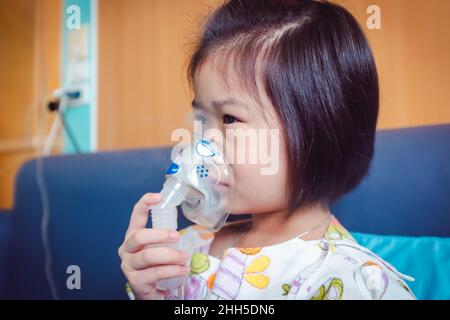 Trauriges asiatisches Kind hält einen Maskendampf-Inhalator zur Behandlung von Asthma. Atmung durch einen Dampfvernebler.Krankheit Mädchen im Krankenhaus eingeliefert. Stockfoto