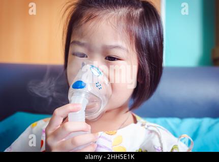 Trauriges asiatisches Kind hält einen Maskendampf-Inhalator zur Behandlung von Asthma. Atmung durch einen Dampfvernebler.Krankheit Mädchen im Krankenhaus eingeliefert. Stockfoto