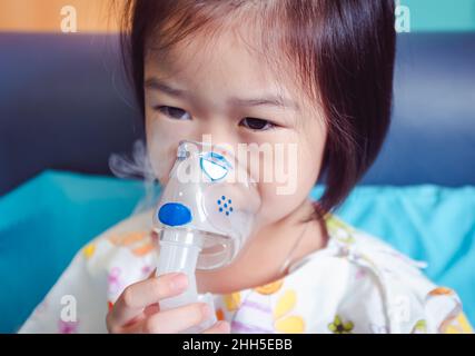 Trauriges asiatisches Kind hält einen Maskendampf-Inhalator zur Behandlung von Asthma. Atmung durch einen Dampfvernebler.Krankheit Mädchen im Krankenhaus eingeliefert. Stockfoto