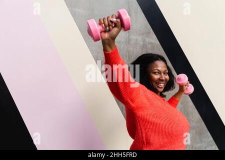 Glückliche Frau mit Hanteln in der Nähe der gemusterten Wand trainieren Stockfoto