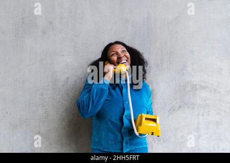 Glückliche Frau, die vor einer grauen Wand mit einem Retro-Telefon spricht Stockfoto