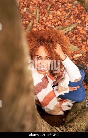 Frau mit der Hand im Haar sitzt am Baum im Herbstwald Stockfoto