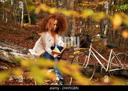 Frau, die im Herbstwald mit dem Fahrrad auf dem Holzbuch sitzt Stockfoto