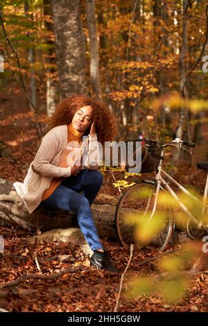 Frau mit der Hand im Haar, die Buch hält, sitzt auf dem Holzbuch mit dem Fahrrad im Herbstwald Stockfoto