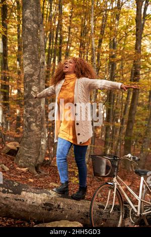 Glückliche Frau mit ausgestreckten Armen, die mit dem Fahrrad im Herbstwald auf dem Baumstamm läuft Stockfoto