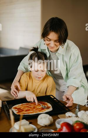 Frau hilft Jungen verteilen Tomatensauce auf Pizzateig in Tablett zu Hause Stockfoto