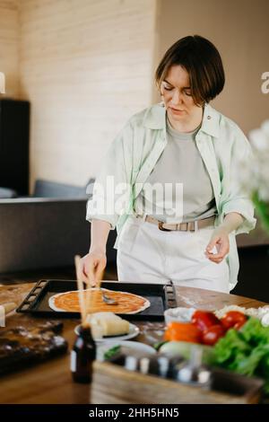 Frau verteilt Sauce auf Teig mit Löffel zu Hause Stockfoto