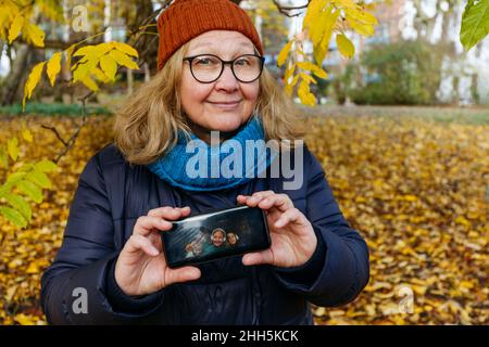 Glückliche ältere Frau, die den Bildschirm des Mobiltelefons mit einem Familienbild im Park zeigt Stockfoto
