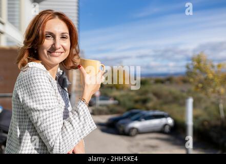 Lächelnde Rothaarige Frau hält den Becher am sonnigen Tag auf dem Balkon Stockfoto