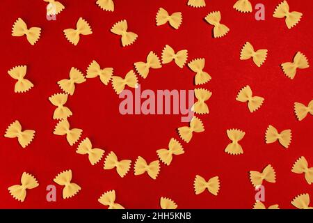 Hintergrund zum Valentinstag. Essen Valentinstag. Pasta-Farfalle-Bögen in Herzform auf rotem Hintergrund zum Valentinstag. Stockfoto