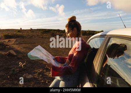 Frau mit Karte, die sich bei Sonnenuntergang auf das Auto stützt Stockfoto