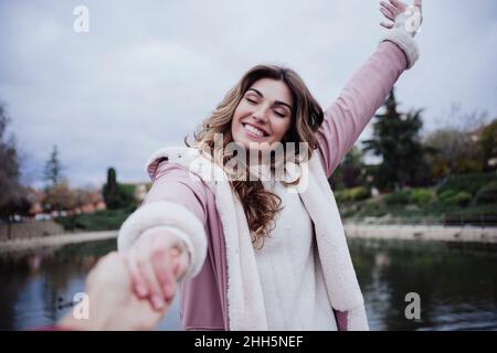 Glückliche Frau, die die Hand des Freundes mit geschlossenen Augen am See hält Stockfoto