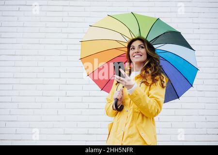 Frau, die Smartphone und Regenschirm vor einer Ziegelwand hält Stockfoto