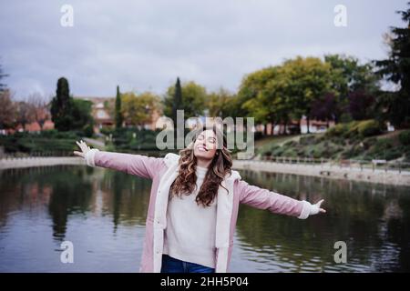 Lächelnde junge Frau mit ausgestreckten Armen am See Stockfoto