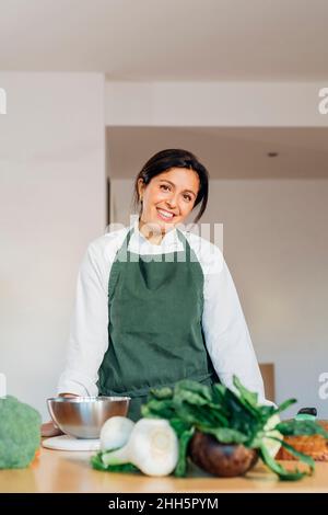Lächelnder Koch mit Schürze, der mit Gemüse am Tisch steht Stockfoto
