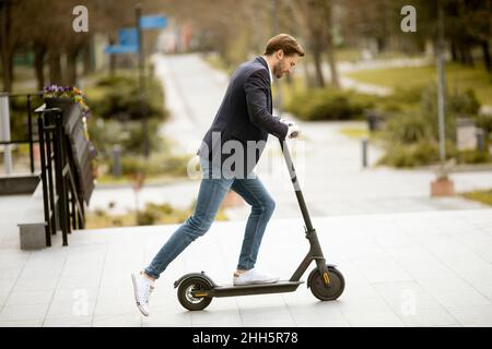 Hübscher junger Geschäftsmann, der auf der Straße durch das Bürogebäude Elektroroller reitet Stockfoto