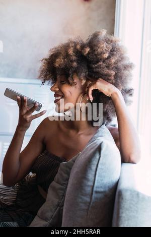 Lächelnde Frau, die zu Hause über das Mobiltelefon Voicemail sendet Stockfoto