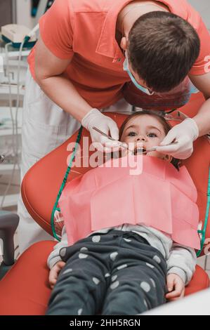 Der Zahnarzt untersucht die Babyzähne des Mädchens, die Behandlung von Babyzähnen, der Zahnarzt hält einen Spiegel in den Händen, Karies bei einem Kind. Stockfoto