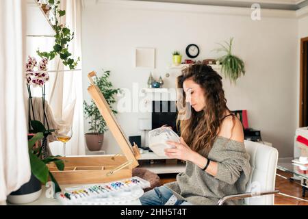 Frau, die Farben mischt, sitzt auf einem Stuhl im Wohnzimmer zu Hause Stockfoto