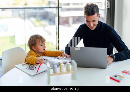 Lächelnder Geschäftsmann, der von seinem Sohn zu Hause auf einem Laptop freiberuflich tätig ist Stockfoto