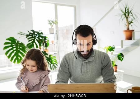 Mann, der ein Headset trägt und einen Laptop verwendet, von der Tochter zu Hause Stockfoto
