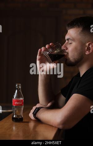 RIVNE, UKRAINE JUNI 26 2020: Junger bärtiger Mann trinkt im Café Coca Cola aus Glas. Flasche mit Cola steht auf einem Holztisch. Stockfoto