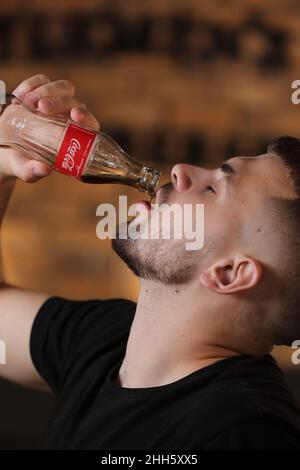 RIVNE, UKRAINE JUNI 26 2020: Junger bärtiger Mann trinkt Coca Cola aus der Flasche im Friseurladen auf Backsteinmauer Hintergrund. Stockfoto