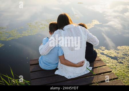Mutter umarmt Söhne, die auf dem Steg vor dem See sitzen Stockfoto