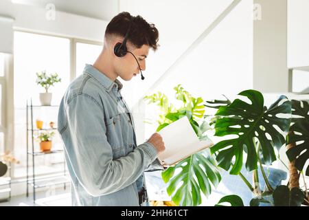 Junger Mann mit Headset liest Tagebuch im Wohnzimmer zu Hause Stockfoto