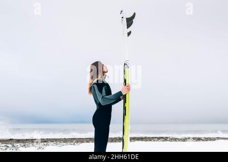 Mann, der im Winter das Surfbrett am Strand anschaut Stockfoto