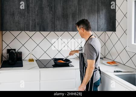 Mann, der Fisch in der Pfanne auf dem Herd zubereitet, zu Hause Stockfoto