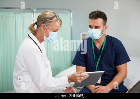 Ärztin mit Gesichtsschutzmaske, die über einen Tablet-PC im medizinischen Raum mit der Krankenschwester diskutiert Stockfoto