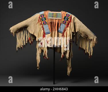 Kriegshemd ca. 1880 Crow, Native American Dieses reich verzierte Kleidungsstück, das Schichten von spiritueller und militärischer Bedeutung verkörpert, wäre bei feierlichen Anlässen von einem Mann von großer Statur getragen worden. Die rote Ockerfarbe auf der oberen Hälfte stellt einen Aspekt heiliger Kraft dar. Die von den Armen und der Brust hängenden gerollten Fransen aus Erminenpelzen zusammen mit den menschlichen Haaren an den Halsklappen bedeuten den Kampf und den Sieg über den Feind. Kriegstrikot. Krähe, Indianer. Ca. 1880. Gegerbtes Leder, Glasperlen, Pigment, Wolltuch, Ermin, Menschliche Haare und Federn. Hergestellt in Montana, Uni Stockfoto