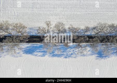 Drohnenansicht des schneebedeckten Feldes und der baumbewachsenen Straße im Winter Stockfoto