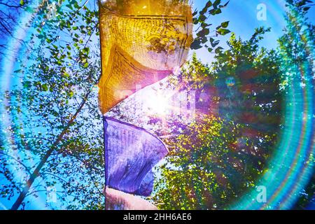 Die bunten buddhistischen Gebetsfahnen, die im Wind gegen den sonnigen Himmel mit bunten Strahlen wiegen, Tibetanischer Garten des Kyiv Botanischen Gartens, Ukraine Stockfoto